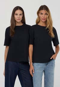 Dwa modele noszą czarne koszulki z okrągłym dekoltem i krótkimi rękawami, zestawione z prostymi dżinsami, prezentując gładki, lekki materiał.