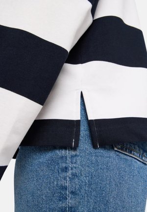 Schwarz-weiß gestreiftes Langarmshirt mit seitlichem Schlitz, getragen über blauen Jeans.