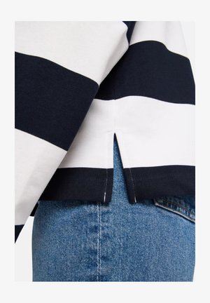 Schwarz-weiß gestreiftes Langarmshirt mit seitlichem Schlitz, getragen über blauen Jeans.