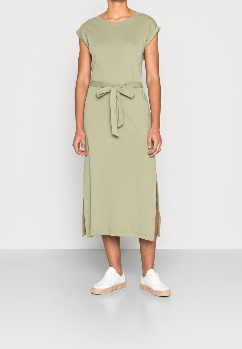 Robe en jersey vert olive à manches courtes, ceinture à la taille, fentes latérales et coupe décontractée, associée à des baskets blanches et une texture lisse.