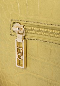 Pelle sintetica verde testurizzata con motivo coccodrillo, dotata di una zip in metallo dorato e dettagli hardware. La zip è parzialmente aperta.