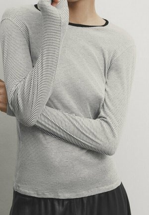 Personne portant une chemise fine à rayures noires et blanches à manches longues avec des trous pour les pouces, bras croisés, et une jupe noire sur un fond uni.