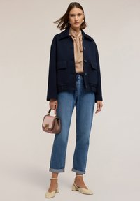 Giacca corta blu navy con tasche, blusa beige, jeans azzurri con risvolto e tacchi color crema. Piccola borsa bicolore con dettaglio logo.