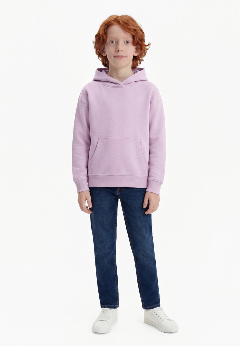 Dítě s červenými vlasy, oblečené v světle fialové mikině, tmavě modrých džínách a bílých teniskách, stojící před jednobarevným bílým pozadím.