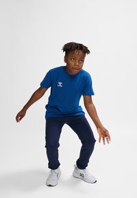 Blauwe katoenen T-shirt met logo, gedragen met marineblauwe sportbroek en witte sneakers. Het kind bevindt zich in een dynamische pose op een lichte achtergrond.