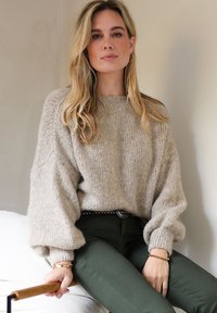 Beige gebreide trui met ballonmouwen, gecombineerd met donkergroene, nauwsluitende broek. De trui heeft een gestructureerde afwerking; een riem met zilveren accenten maakt de look af.