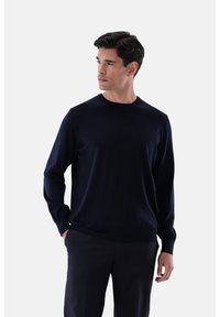 Mann mit dunklen Haaren, der einen navyblauen Langarmpullover und dunkle Hosen trägt, steht mit einer Hand in der Tasche und schaut zur Seite.