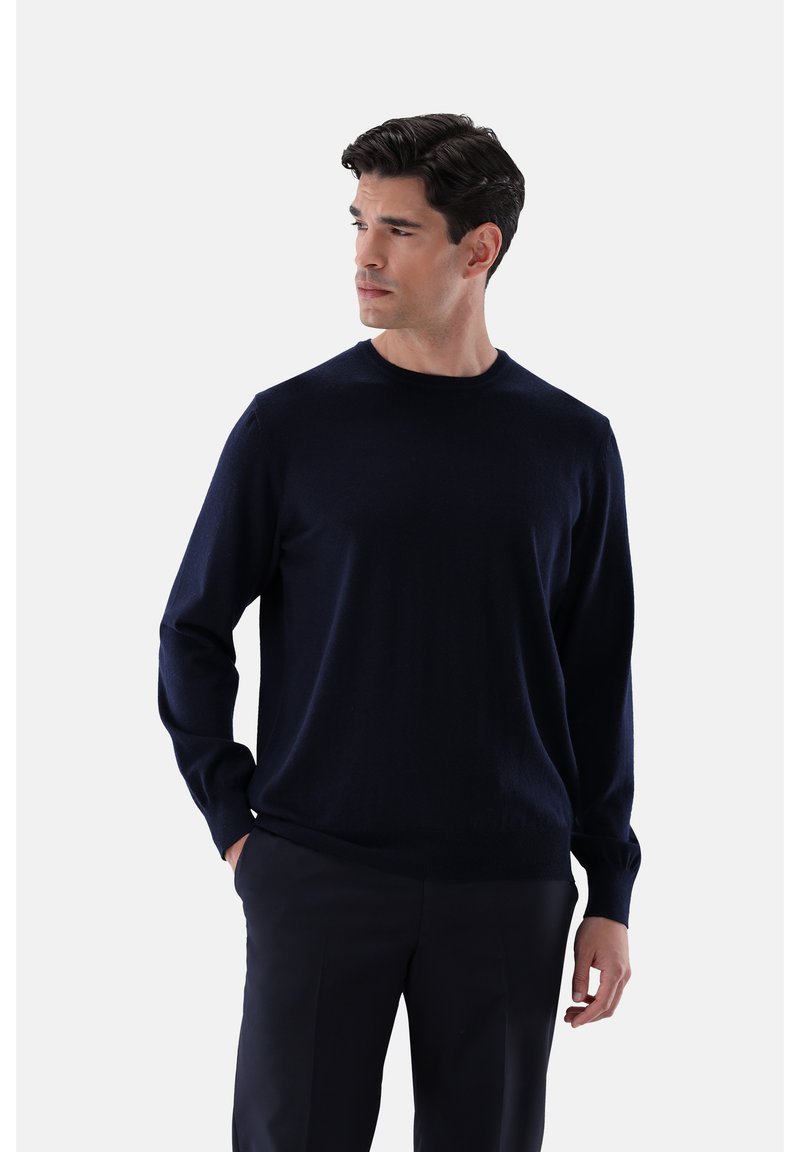 Mann mit dunklen Haaren, der einen navyblauen Langarmpullover und dunkle Hosen trägt, steht mit einer Hand in der Tasche und schaut zur Seite.