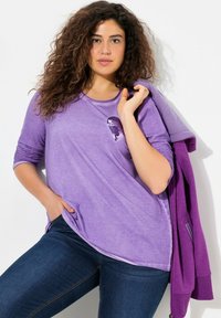 Lila Langarmshirt aus weichem Stoff mit einem Paillettendetail in Herzform. Trägt dazu eine dunkelblaue Jeans, der Model posiert lässig.