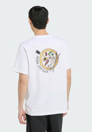 Muž nosící bílé tričko Adidas s barevným motivem sushi a hůlek a nápisem „Adidas Sportswear“ na zádech.