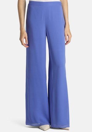 Donna che indossa pantaloni viola a vita alta e a gamba larga abbinati a scarpe beige con punta, vista dalla vita ai piedi su sfondo bianco.