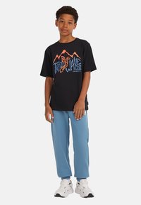 Zwarte katoenen T-shirt met een kleurrafisch ontwerp van bergen en tekst. Lichtblauwe sweatpants en witte sneakers maken de outfit compleet.