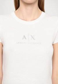Maglietta in cotone bianco con scollatura rotonda, maniche corte e logo in paillettes argento "A|X ARMANI EXCHANGE" sul davanti.