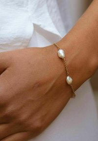 Goldene Kettenarmband mit zwei unregelmäßig geformten weißen Perlen, die auf einem gebräunten Handgelenk ruhen, vor einem hellen Stoffhintergrund.