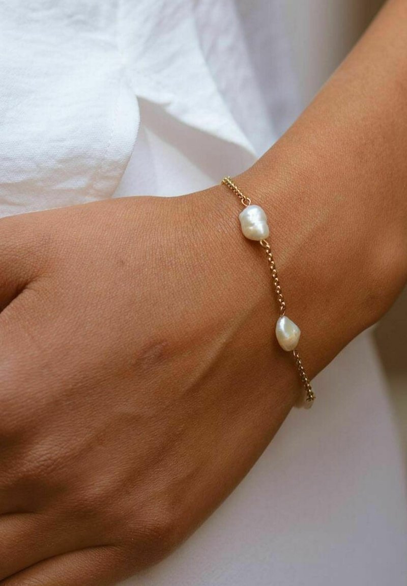 Goldene Kettenarmband mit zwei unregelmäßig geformten weißen Perlen, die auf einem gebräunten Handgelenk ruhen, vor einem hellen Stoffhintergrund.