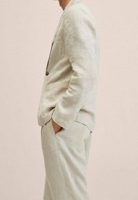 Jasny lniany garnitur w luźnym kroju, z zapięciem na guzik, bocznymi kieszeniami i gładką teksturą. Stylizowany z rękami w kieszeniach.