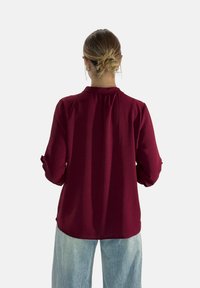 Blusa bordeaux a maniche lunghe con una finitura testurizzata, polsini abbottonati e colletto, abbinata a jeans azzurri.