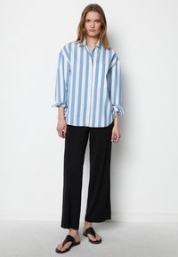 Camicia button-up a righe blu chiaro e bianco con taschino; abbinata a pantaloni neri larghi e sandali neri. Tessuto morbido e vestibilità rilassata.