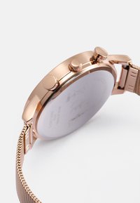 Rose-guldfarvet metalur med mesh-bånd, rund størrelse, tre sideknapper og et glasbagbeklædning, der viser et svagt design.