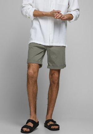 Man som bär uppvikta olivgröna shorts, vit långärmad skjorta och svarta remsandaler, stående mot en grå bakgrund.