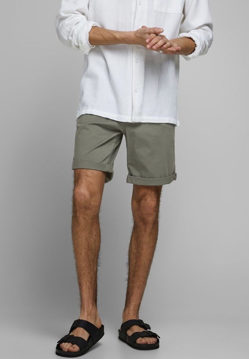 Uomo con pantaloni corti verde oliva arrotolati, camicia bianca a maniche lunghe e sandali neri con cinturino, in piedi su uno sfondo grigio.