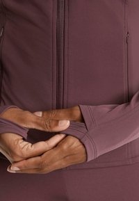 Veste de sport mauve avec une texture lisse, fermeture éclair à l'avant et trous pour les pouces ; gros plan des mains ajustant le tissu au niveau des poignets.