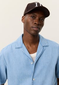 Ανοιχτό μπλε πουκάμισο με κοντά μανίκια, σχεδιασμό με γιακά, υφή υφάσματος και διακριτική λεπτομέρεια λογότυπου. Φορεμένο πάνω από λευκό сет романτικά τοπ. Καφέ καπέλο.