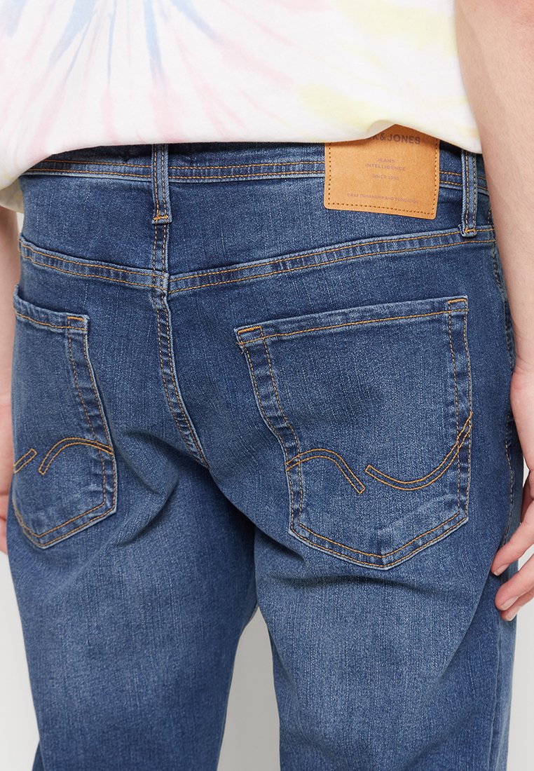 Jeans de mezclilla azul oscuro con textura suave, costuras naranjas y dos bolsillos traseros, con una etiqueta de cuero marrón en la cintura.