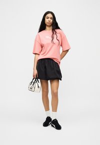 Jeune femme aux longs cheveux portant un t-shirt rose oversize, une jupe noire, des chaussettes blanches, des baskets noires, tenant un petit sac à main blanc Puma.