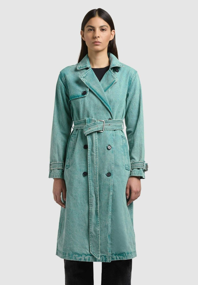 Donna che indossa un lungo trench in denim doppio petto color verde acqua sbiadito con cintura e pantaloni neri, in piedi davanti a uno sfondo chiaro.