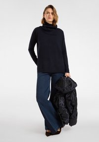 Maglione a collo alto in maglia blu navy, vestibilità comoda; abbinato a jeans blu a zampa. Il modello tiene in mano una giacca imbottita nera con motivo trapuntato.