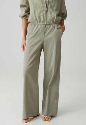 Vrouw draagt lichtolijfgroene broek met wijd uitlopende pijpen, elastische taille en zijzakken, gecombineerd met beige sandalen met vierkante neus.