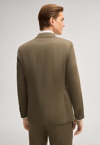 Mann von hinten, trägt eine olivgrüne Anzugjacke und passende Hose vor einem schlichten hellen Hintergrund.