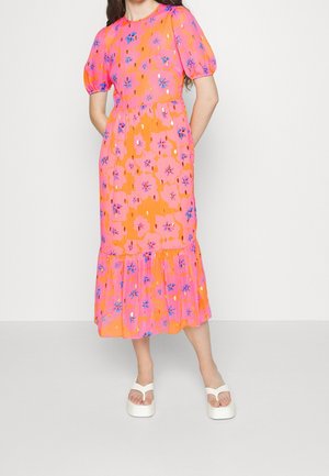 Mujer vistiendo un vestido midi floral de colores naranja brillante y rosa con mangas abullonadas y sandalias blancas de plataforma, de pie con las manos en los bolsillos.
