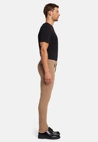 Lichtbruine slim-fit broek gecombineerd met een zwart t-shirt en zwarte schoenen, weergegeven in een zijprofiel tegen een effen achtergrond.