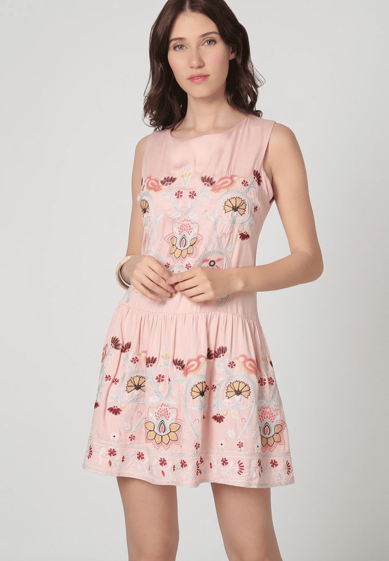 Mujer con un vestido rosa sin mangas con bordados florales, de pie contra un fondo claro y liso, con las manos entrelazadas al frente.