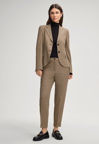 Completo marrone a quadratini con giacca aderente e pantaloni tapered, abbinato a un dolcevita nero a coste e mocassini neri. Texture liscia.