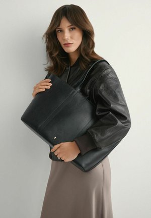 Kobieta o brązowych włosach, ubrana w czarną skórzaną kurtkę i taupe'ową spódnicę, trzymająca dużą czarną skórzaną torbę typu tote na neutralnym tle.