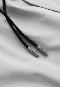 Cordonnettes tressées noires pour sweat à capuche avec embouts métalliques gravés du mot "ombre" sur fond en tissu gris clair.