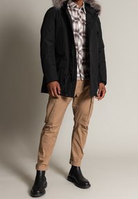 Svart isolerad parkas med faux päls huva, i kombination med en rutig skjorta och beiga cargo-byxor. Modellen har på sig svarta ankelstövlar.