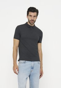 Άνδρας με σκούρο γκρι κοντομάνικο πόλο και ανοιχτά μπλε τζιν, στέκεται μπροστά σε λευκό φόντο, κοιτάζοντας μπροστά.