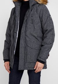 Parka gris acolchada con capucha de pelo sintético, bolsillos con cremallera y puños acanalados. Tejido texturizado con un patrón sutil.