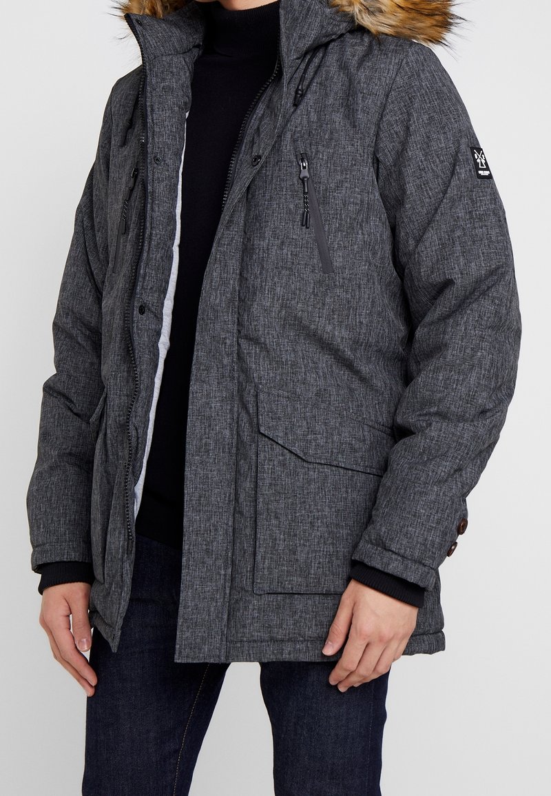 Parka gris acolchada con capucha de pelo sintético, bolsillos con cremallera y puños acanalados. Tejido texturizado con un patrón sutil.