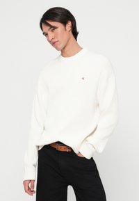Pulover alb cu dungi, cu guler rotund, mâneci lungi și un mic logo roșu pe piept, asociat cu pantaloni negri și o curea maro.