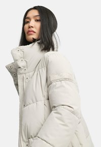 Chaqueta tipo plumón en gris claro, con un alto cuello, detalles de botones y un parche texturizado en la manga. Tejido suave y acolchado.