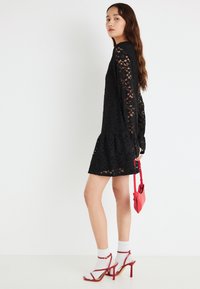 Μαύρο φόρεμα από δαντέλα με μακριά μανίκια και φούστα σε στρώσεις. Συνδυάζεται με κόκκινες σανδάλες με λουράκια και μικρή κόκκινη τσάντα χειρός. Ορατές λευκές κάλτσες.