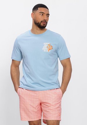 Férfi világoskék pólóban, amelyen kis korall grafikája van, és rózsaszín mintás rövidnadrágban áll, kezét zsebében tartva, fehér háttér előtt.
