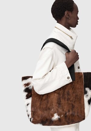 Persona con una giacca bianca che porta una grande borsa a tracolla con motivo in pelle di mucca marrone e bianca.