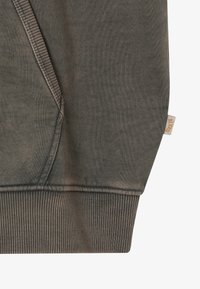 Zbliżenie na zieloną bluzę z kapturem z kieszenią, z ryflowanym dołem i małą beżową metką z napisem "BLKINS" po prawej stronie.