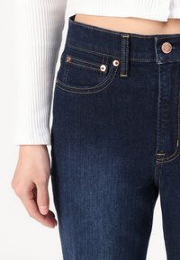 Jeans in denim blu scuro con un design aderente, cuciture arancioni e stile a cinque tasche. Include accessori in tonalità rame e chiusura con bottone.